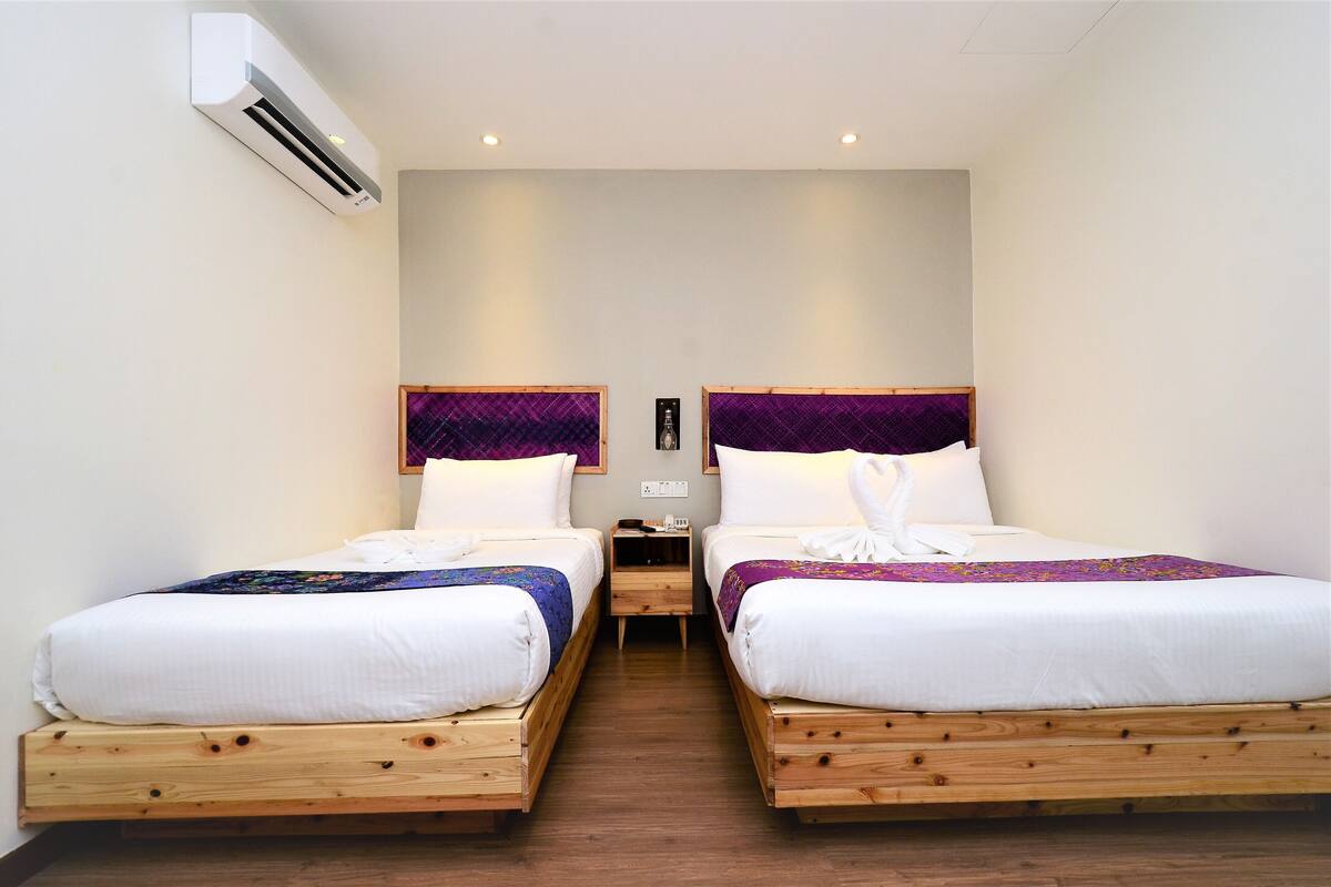 A hotel room with two wooden beds, white bedding, purple headboards, and an air conditioning unit on the wall.