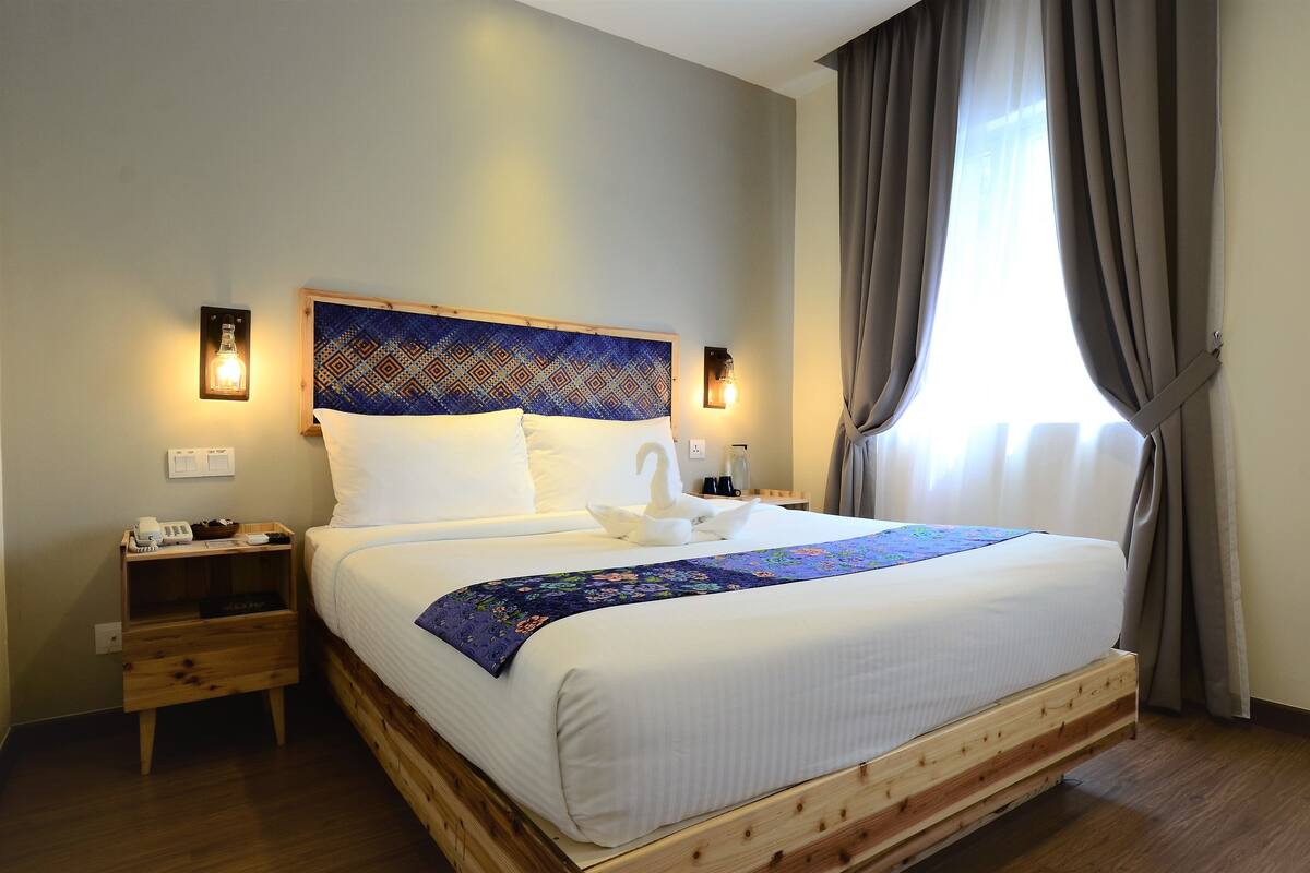 A neatly made bed with a swan-shaped pillow and a blue floral bedspread. There are two bedside tables with lamps, a wooden nightstand, and a window with curtains.
