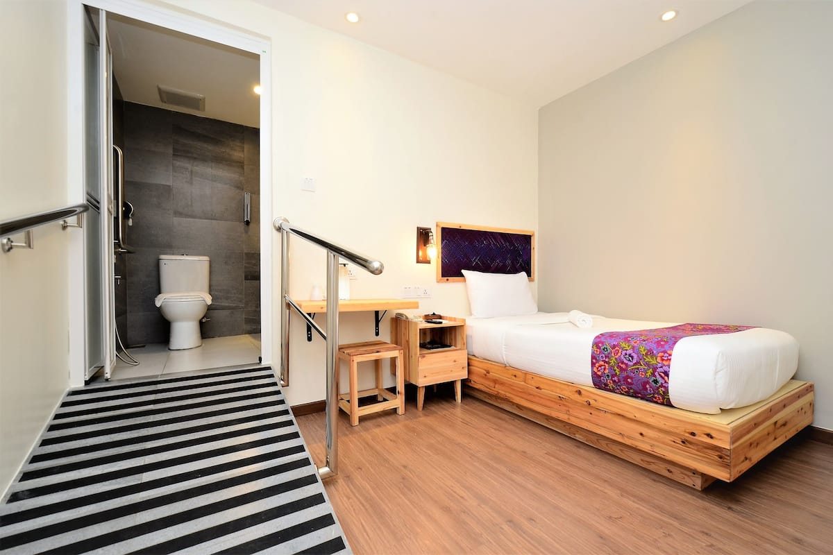 A compact bedroom with a wooden bed, a bedside table, a nightstand, a small stool, and a bathroom with a toilet and shower.