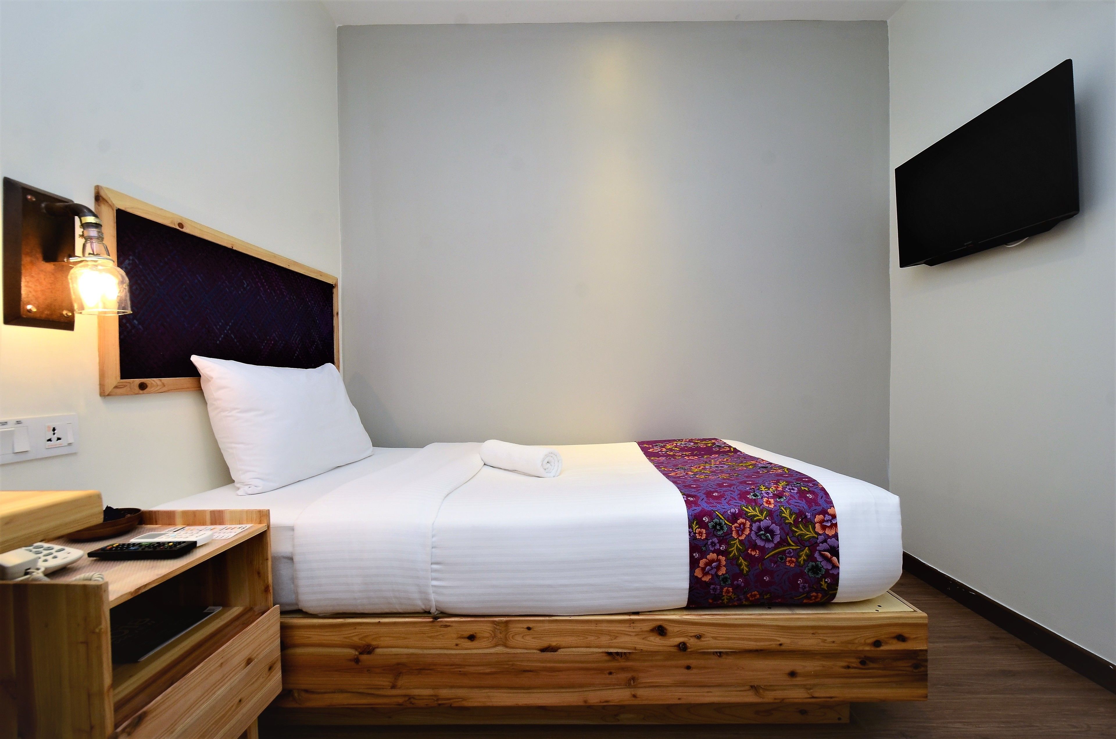 A single bed with a white mattress and a colorful floral bedspread, a wooden nightstand with a remote control, and a wall-mounted TV.