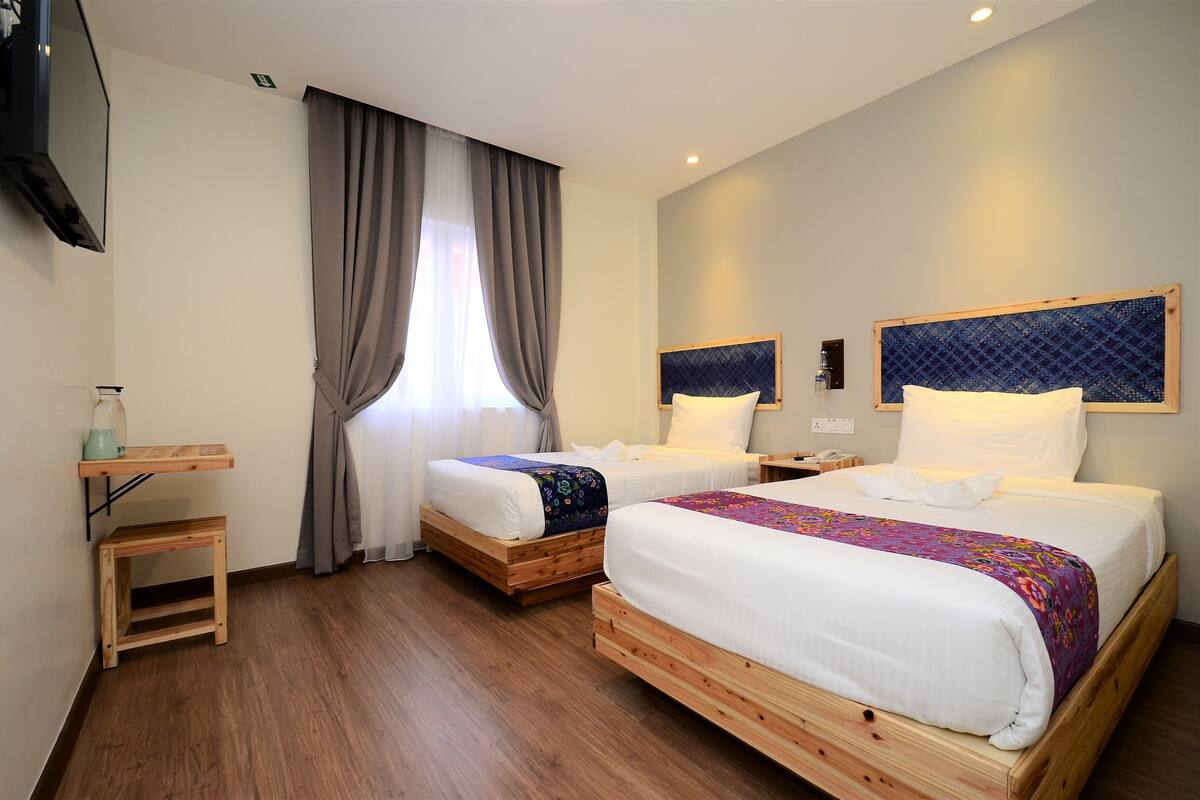 A hotel room with two wooden beds, a small wooden table, a TV mounted on the wall, and a window with curtains.
