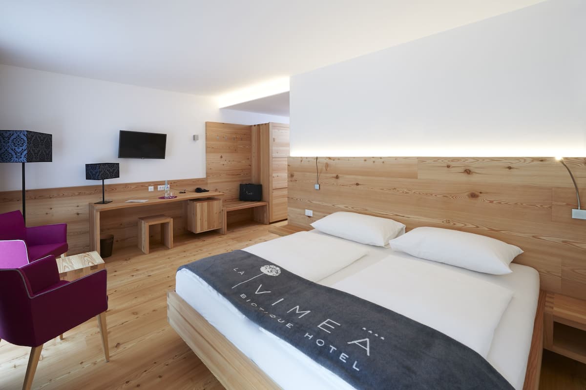A modern hotel room with a wooden headboard, a bed with white linens, a desk with a chair, a flat-screen TV, and a large window with curtains.