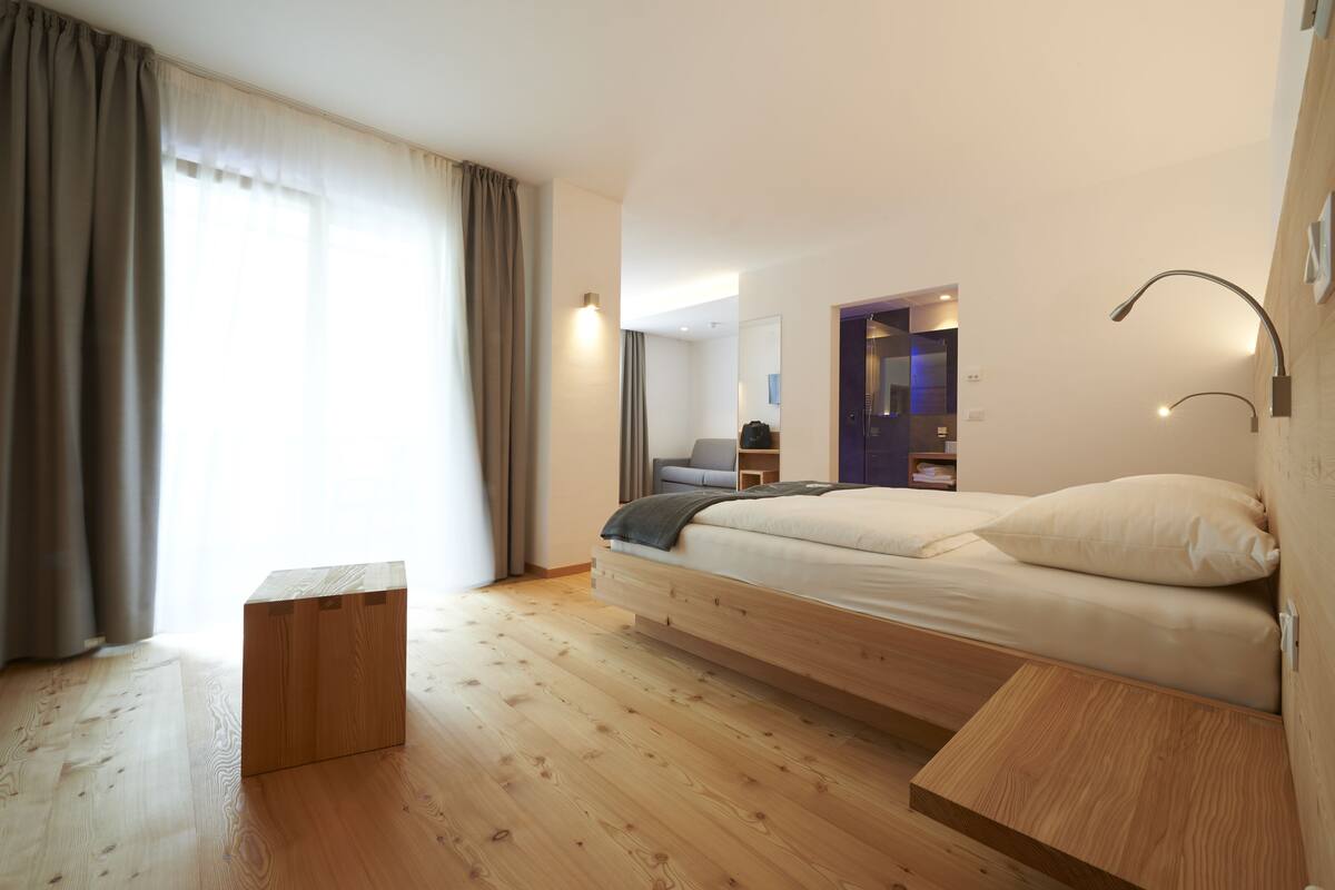 A modern bedroom with a wooden floor, a bed with a headboard, a small wooden nightstand, a nightstand lamp, and a large window with curtains.