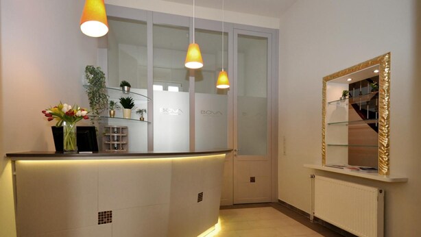 A reception area with a white reception desk, two pendant lights, a glass display cabinet, and a mirror with a gold frame. - Bova Hotel Frankfurt (Frankfurt)