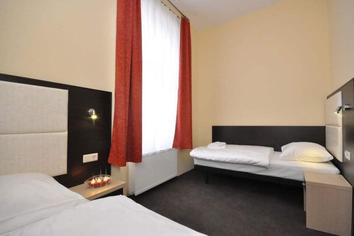 Quarto de hotel com duas camas, uma mesinha de cabeceira com uma vela, uma luminária na parede e uma janela com cortinas vermelhas.