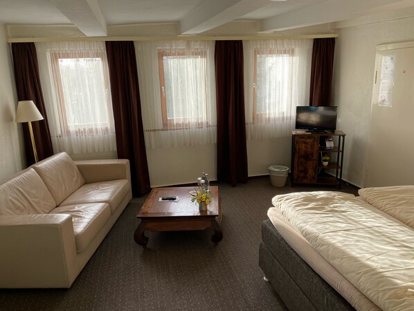 A hotel room with a sofa, a bed, a TV on a wooden stand, and a window with curtains.
