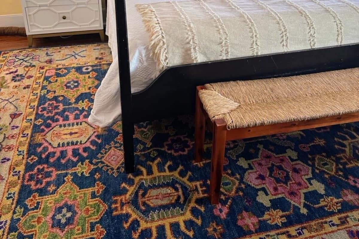 Un lit à baldaquin avec une couvre-lit blanche et beige, un coussin coloré et un banc en bois au pied du lit.