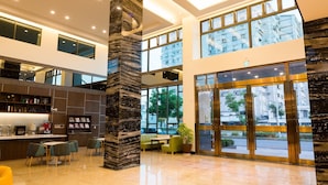 Een moderne hotellobby met een hoge plafond, grote ramen, een centrale pilaar, een zithoek en een receptiebalie.