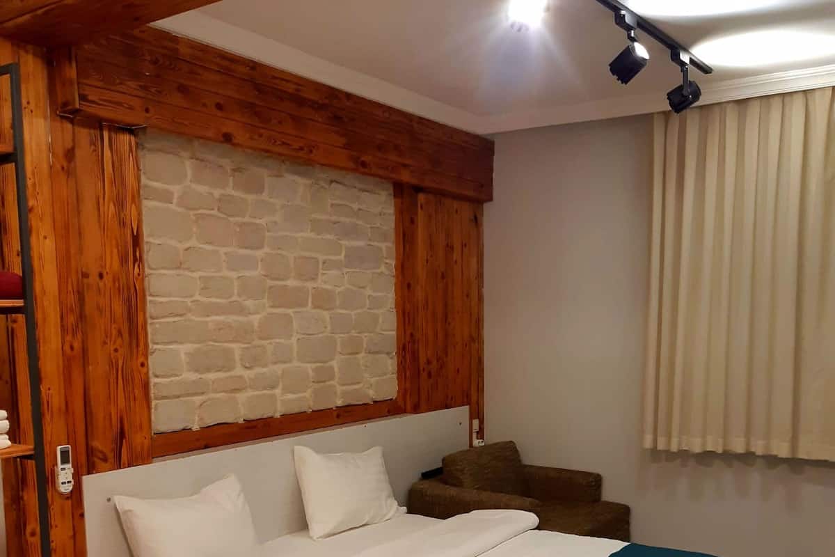 A bedroom with a wooden ceiling, a brick wall, a bed with white bedding and a blue blanket, a wooden bench, and a window with curtains.