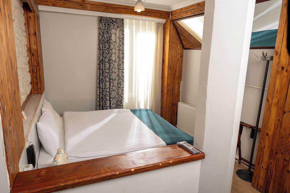 A bedroom with a wooden headboard, a bed with white bedding, a wooden bedside table, and a window with sheer curtains.