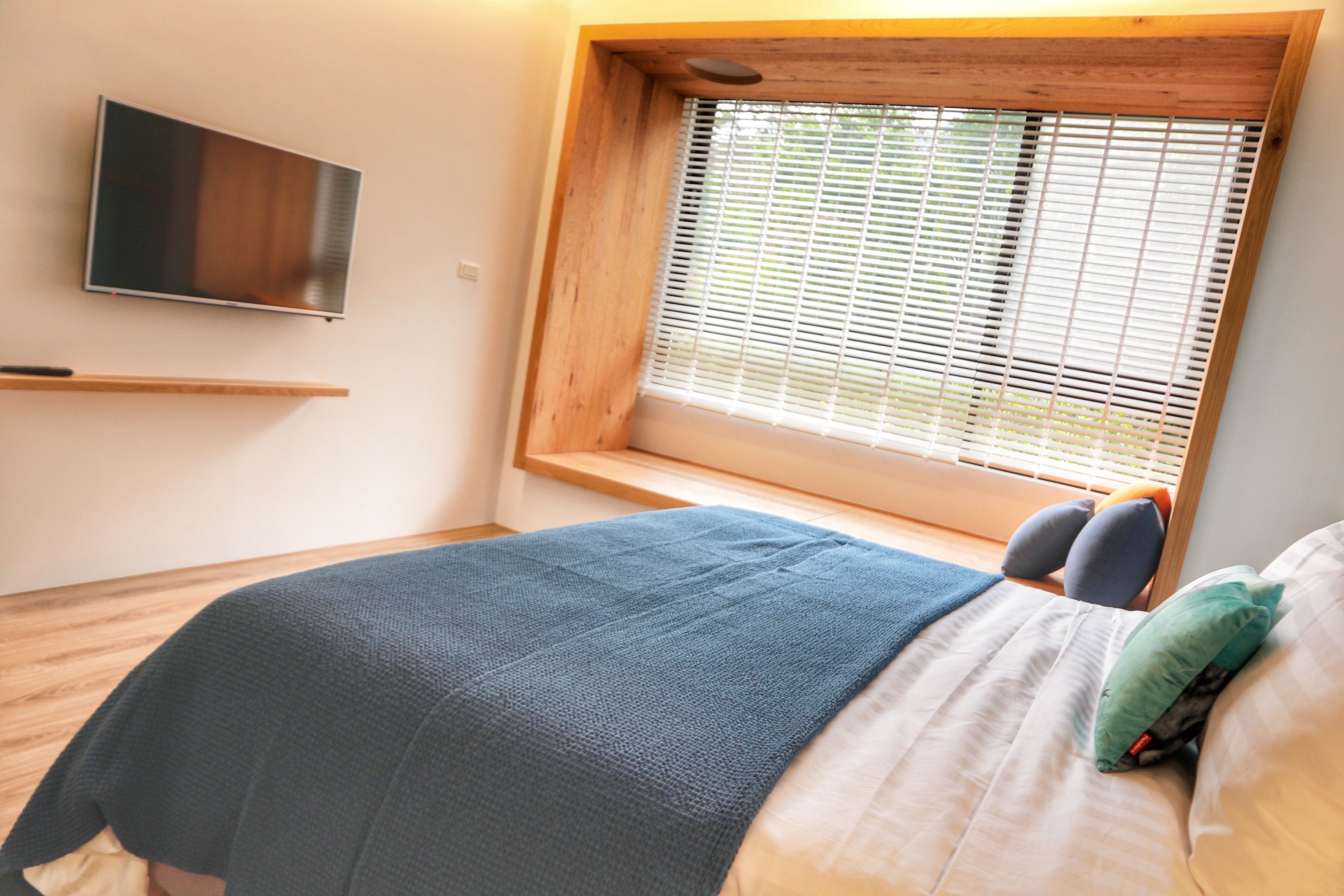 A bedroom with a bed, a TV mounted on the wall, a window with blinds, and a wooden shelf.