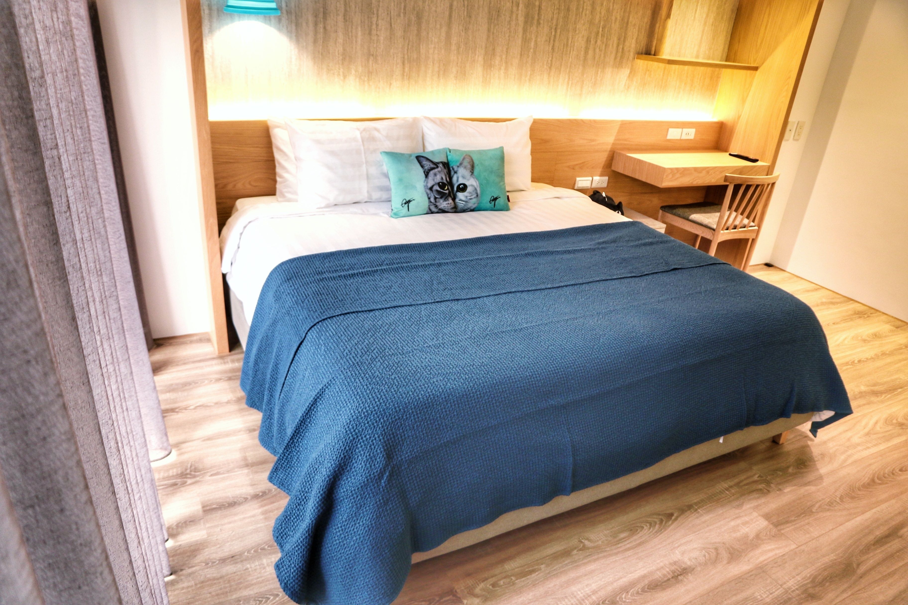 A neatly made bed with a blue blanket, a pillow with a cat design, and a wooden headboard.