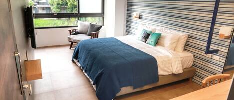 A modern bedroom with a large bed, a desk, and a window with a view of greenery outside.