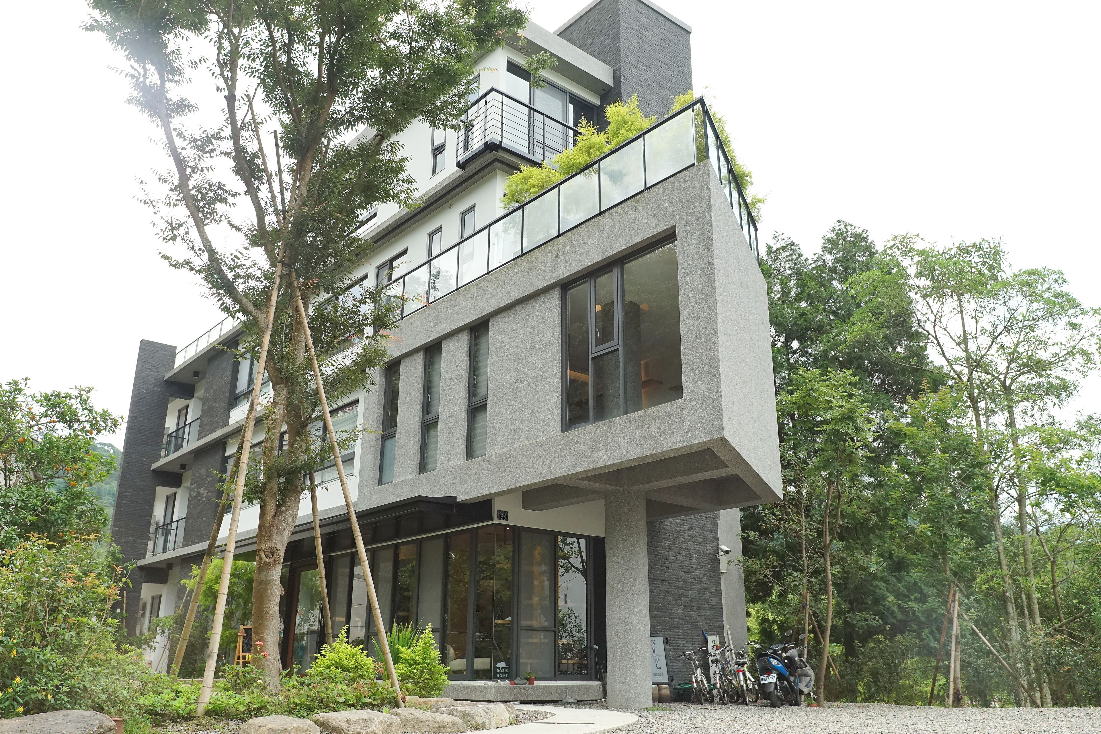 A modern multi-story building with large windows, a balcony with greenery, and a gravel driveway.
