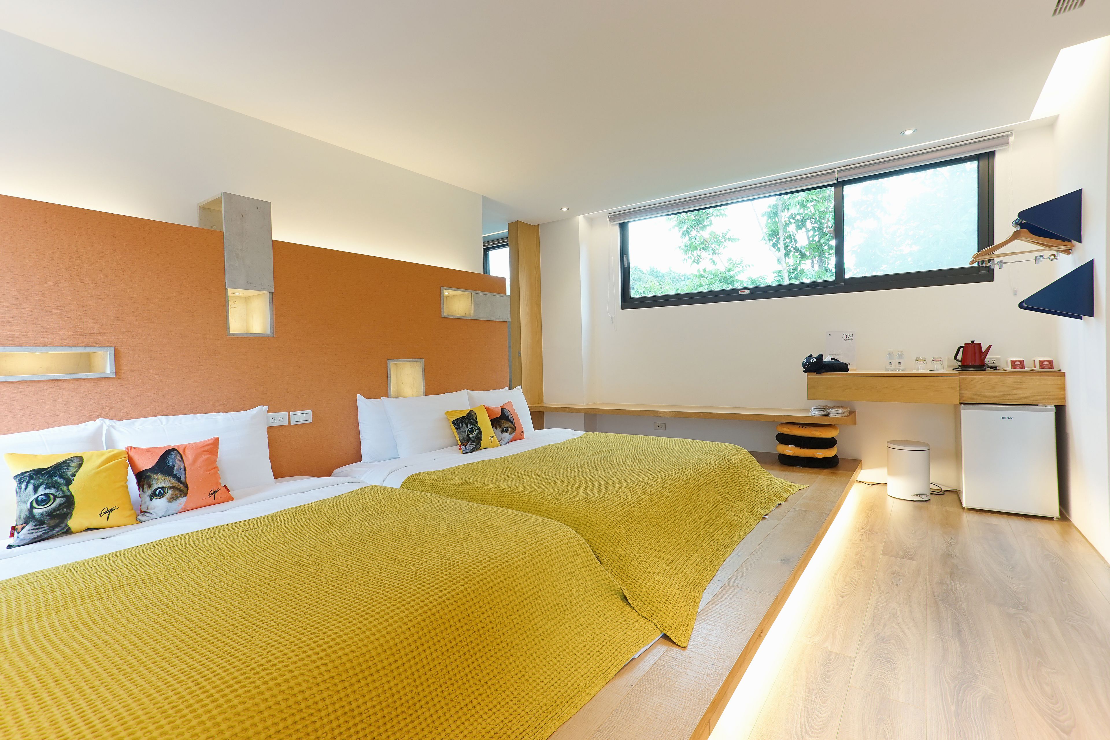 Una habitación de hotel moderna con dos camas, un escritorio, un refrigerador compacto y un ventanal grande que proporciona luz natural.