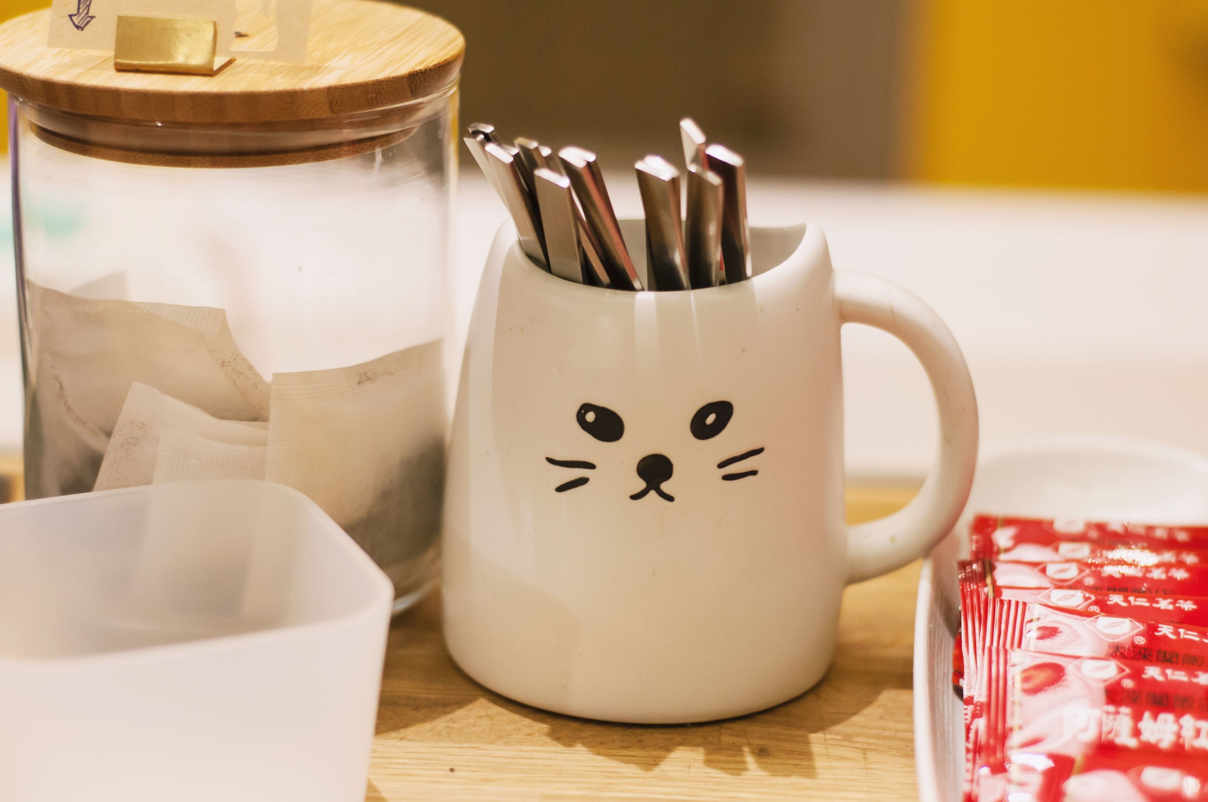 A white mug with a cat face design, a jar with a wooden lid, and a red packet on a wooden surface.