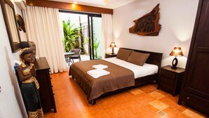 A hotel room with a bed, two bedside tables, a wall-mounted artwork, a statue, and a sliding glass door to a balcony with plants. - Penn Sunset Villa 4 (Ko Lanta)