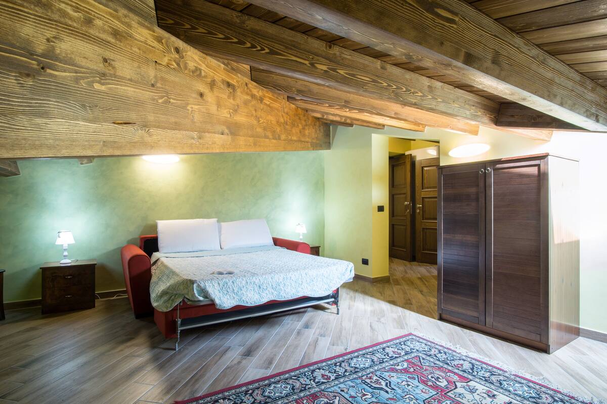 Een slaapkamer met een bed, houten balken in het plafond, een nachtkastje, een garderobe en een kamerkleed met een patroon.