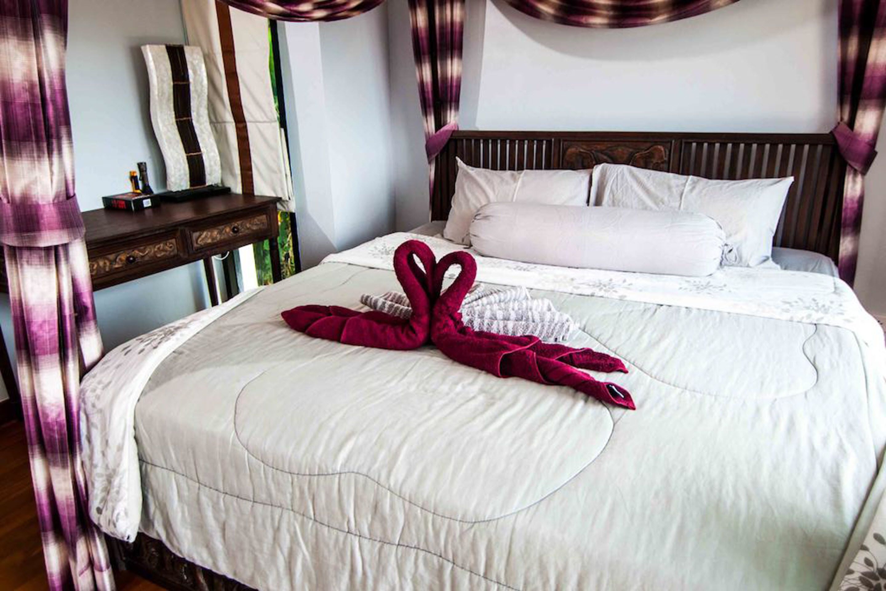 A neatly made bed with a heart-shaped towel arrangement, a wooden nightstand with a mirror, and a headboard with vertical slats.