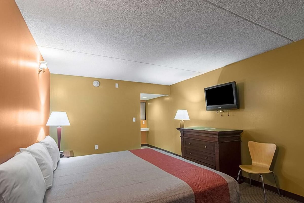 A hotel room with a bed, two bedside lamps, a flat-screen TV mounted on the wall, a wooden dresser, and a chair.