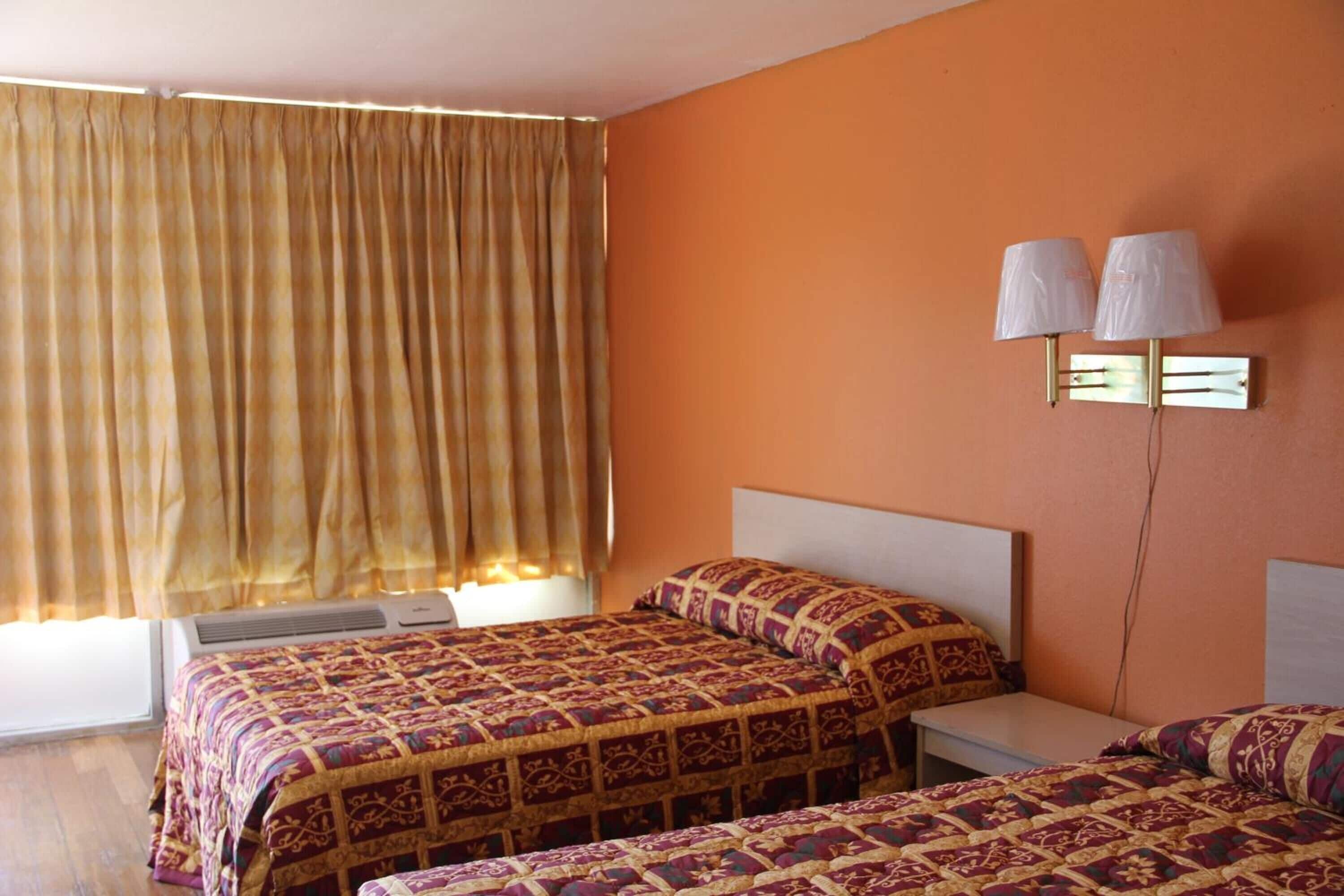 A hotel room with two beds, a window with curtains, an air conditioning unit, and a wall-mounted lamp.