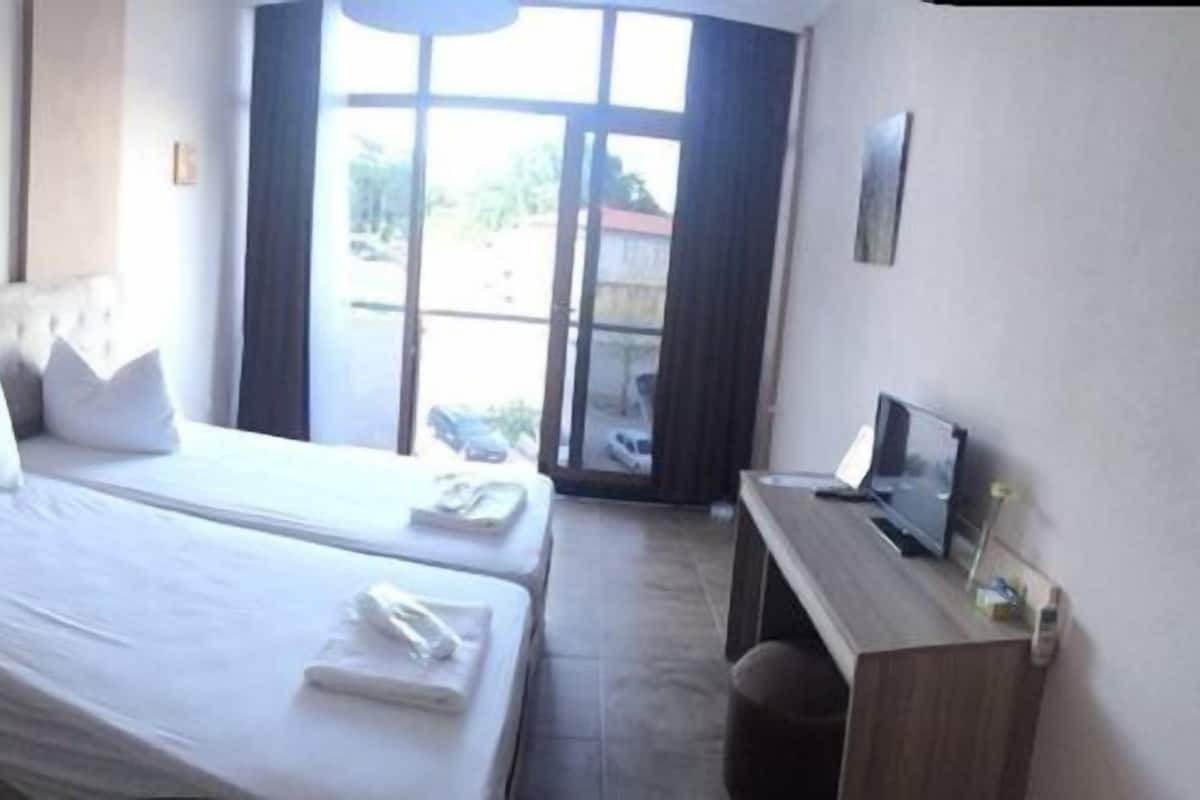 A bedroom with two beds, a bedside table, a desk, and a window with a view.