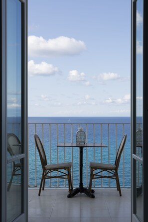 Un balcón con dos sillones de mimbre y una mesita pequeña, con vista a un mar y cielo azul despejado.