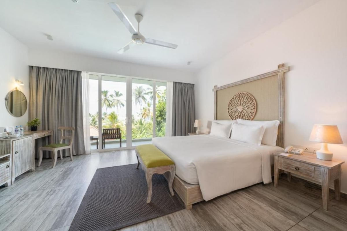 A spacious bedroom with a large bed, a desk, a chair, a small table, and a nightstand with a lamp.