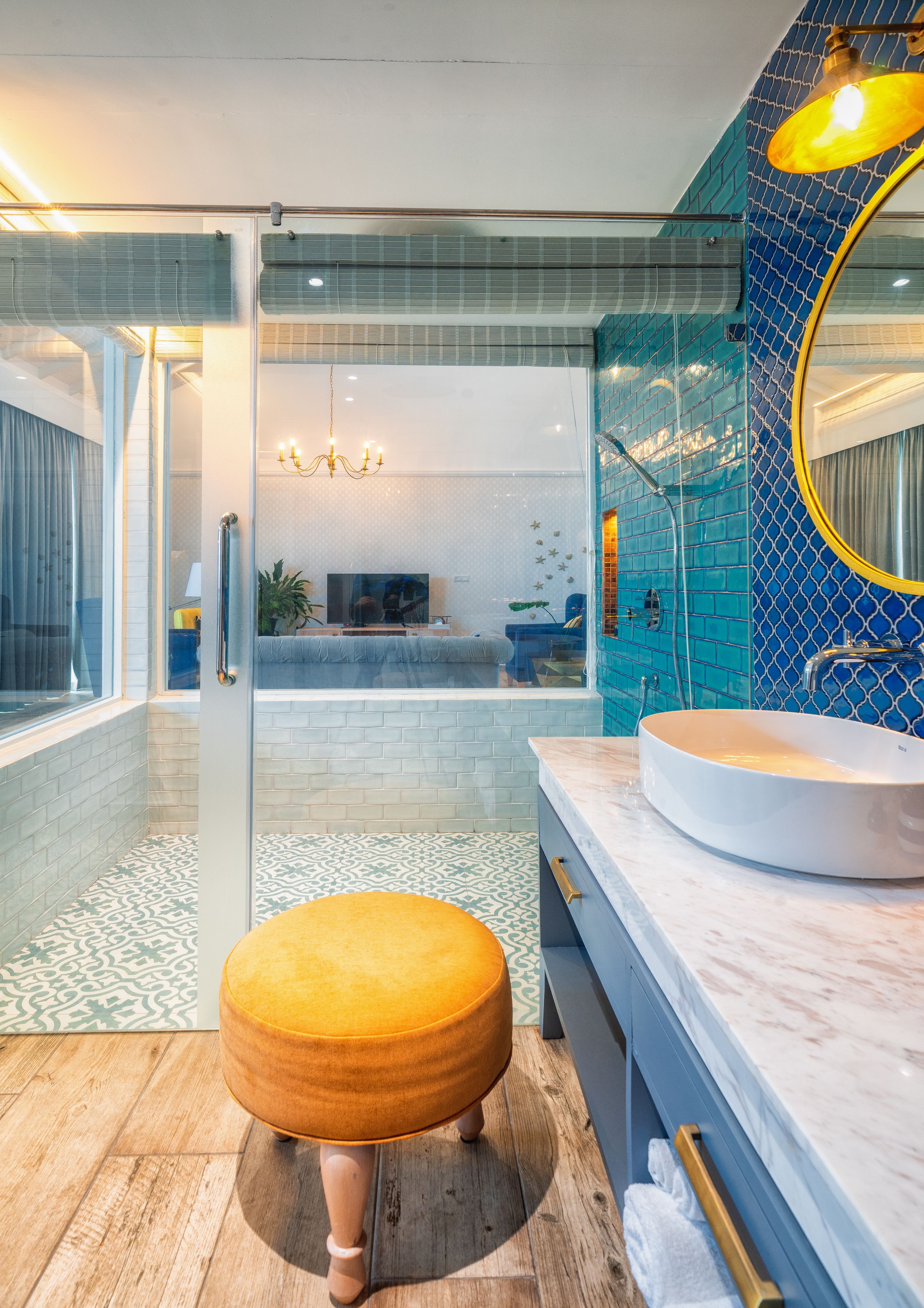 A modern bathroom with a glass shower enclosure, a round yellow stool, a white sink, and a blue-tiled wall.