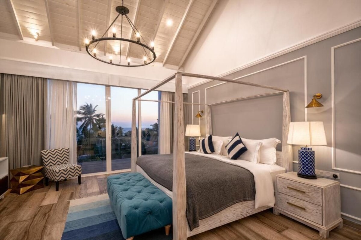 A bedroom with a four-poster bed, a large window with a view, a chandelier, and a nightstand with a lamp.