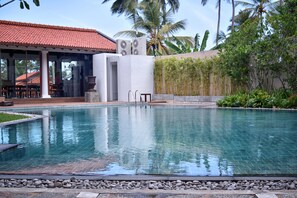 A swimming pool with a tiled edge, a covered seating area with chairs, and a building with a red-tiled roof.