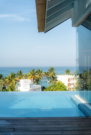A clear view of a swimming pool with a wooden deck, overlooking a beach with palm trees and a white building in the background.
