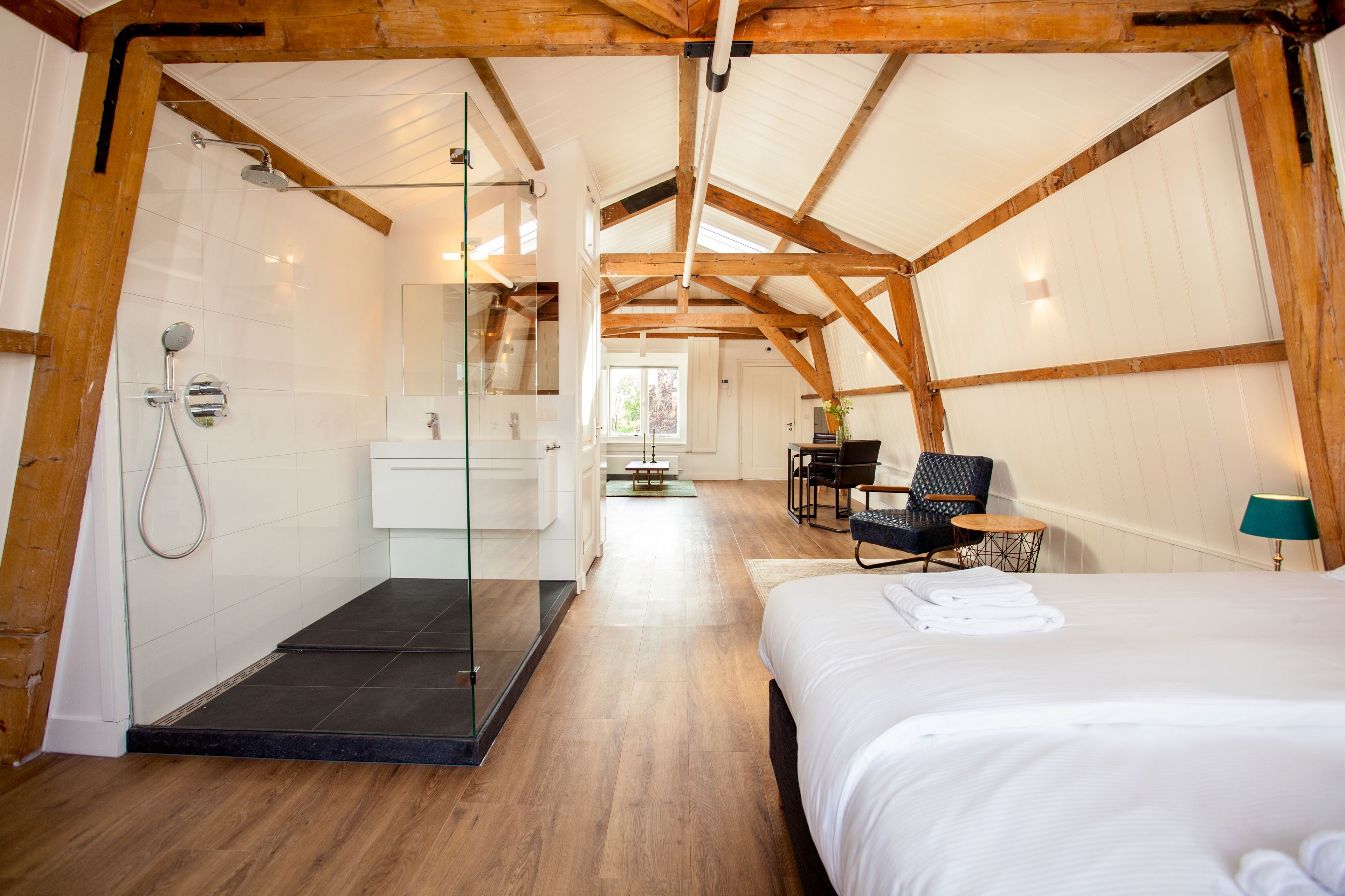 Een moderne hotelkamer met een houten plafond, een glazen douchecabine, een bed met netjes opgevouwen handdoeken en een zithoek met een stoel en tafel.