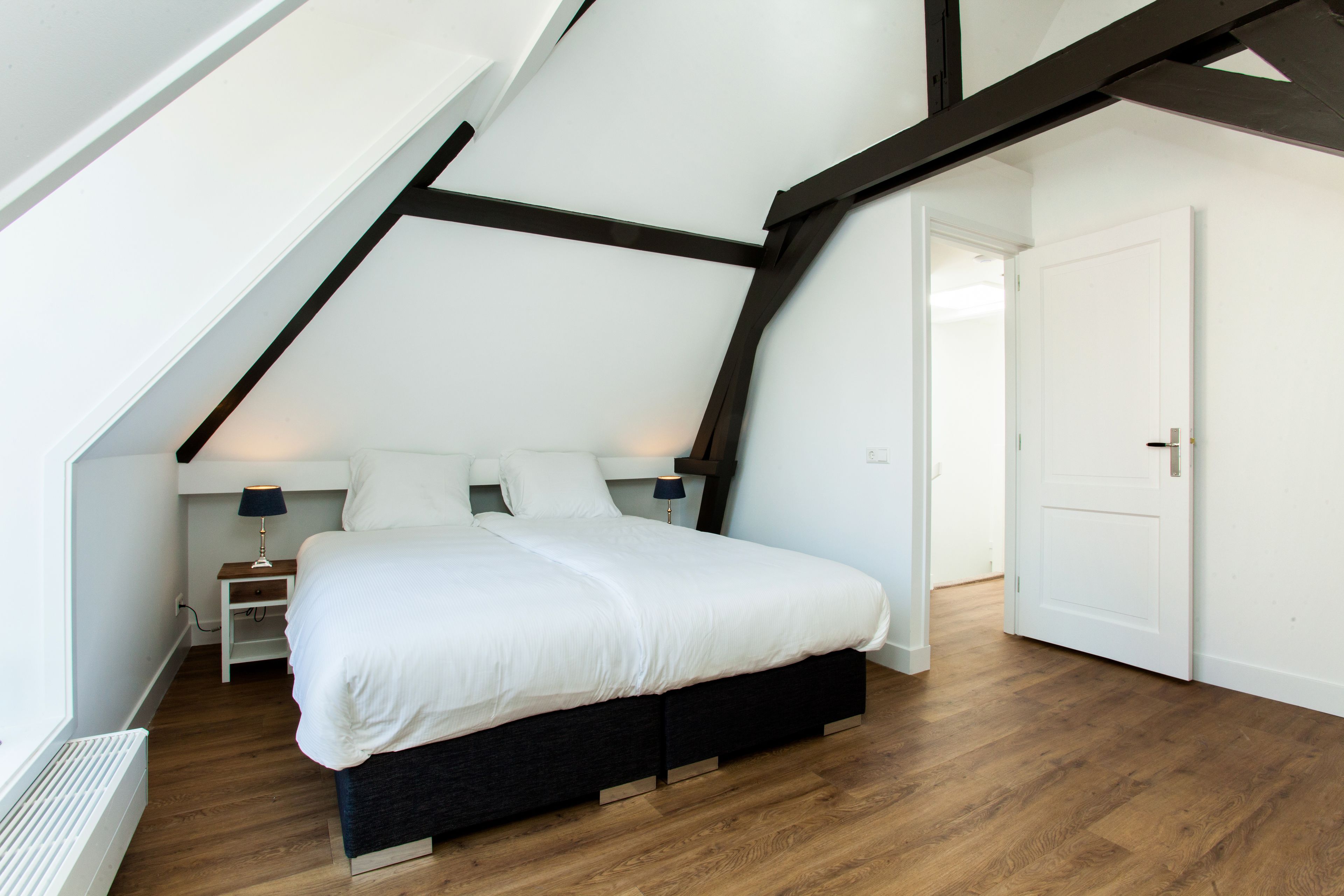 Ein Schlafzimmer mit schrägen Wänden, einem Bett mit weißer Bettwäsche, einem Nachttisch mit Lampe und Holzboden.