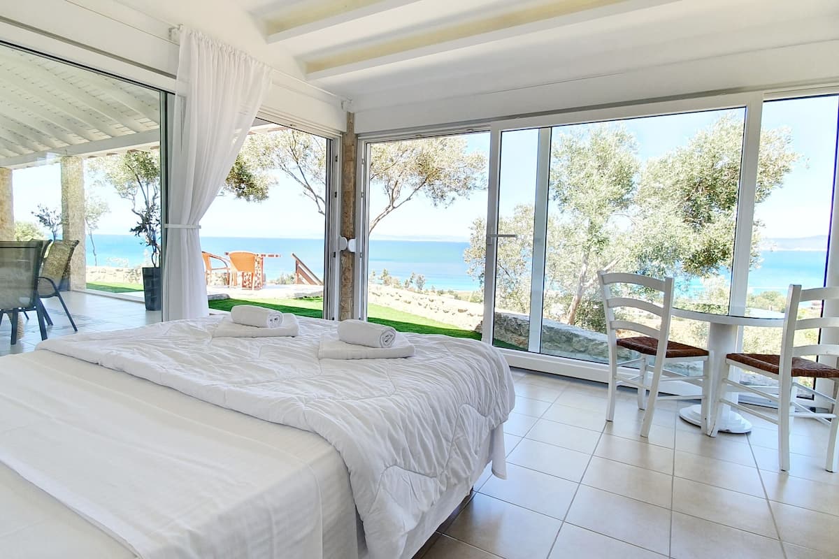 Une chambre dotée d’un grand lit, d’une porte vitrée donnant sur un balcon avec vue sur la mer, et d’un coin repas avec des chaises et une table.