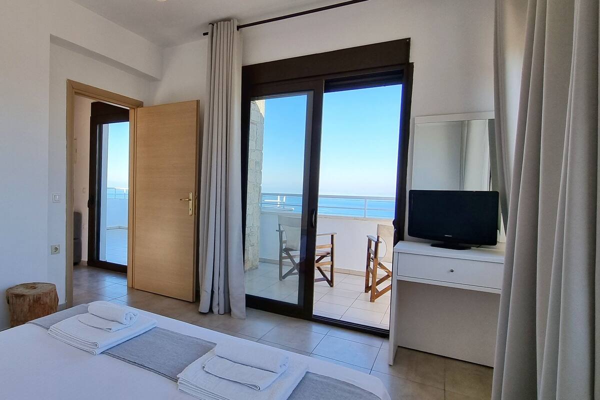 Une chambre d’hôtel moderne avec un grand lit, un téléviseur posé sur une table d’appoint et un balcon offrant une vue sur la mer.