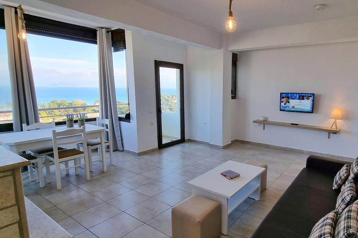 Un salon moderne avec un canapé, une table basse, un téléviseur fixé au mur et un balcon offrant une vue sur la mer.