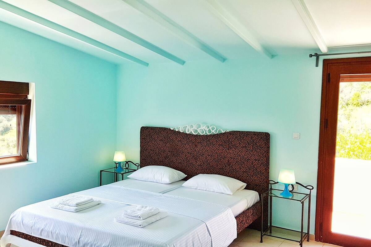 Une chambre à coucher avec un lit, deux tables de chevet avec des lampes, une fenêtre avec des rideaux et une vue sur la verdure à l’extérieur.