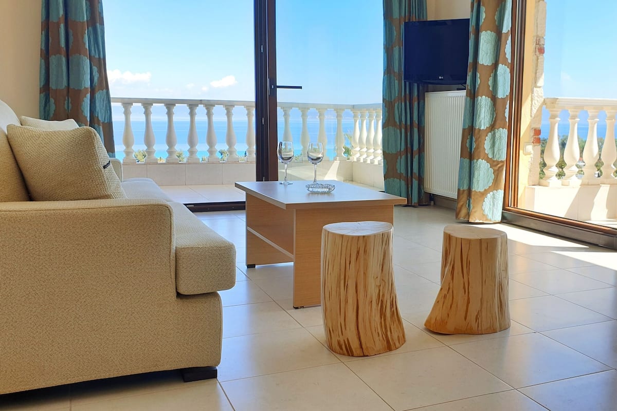 Un salon comprenant un canapé, une table basse et deux tabourets en bois. Un téléviseur est fixé au mur, et il y a un balcon avec une balustrade et une vue sur la mer.
