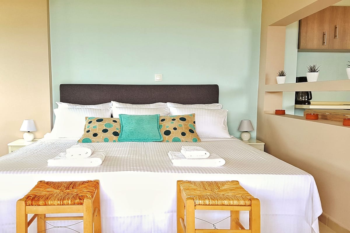Un lit bien fait, avec du linge de lit blanc, une tête de lit sombre et des coussins décoratifs. On peut voir deux tabourets en bois devant le lit.