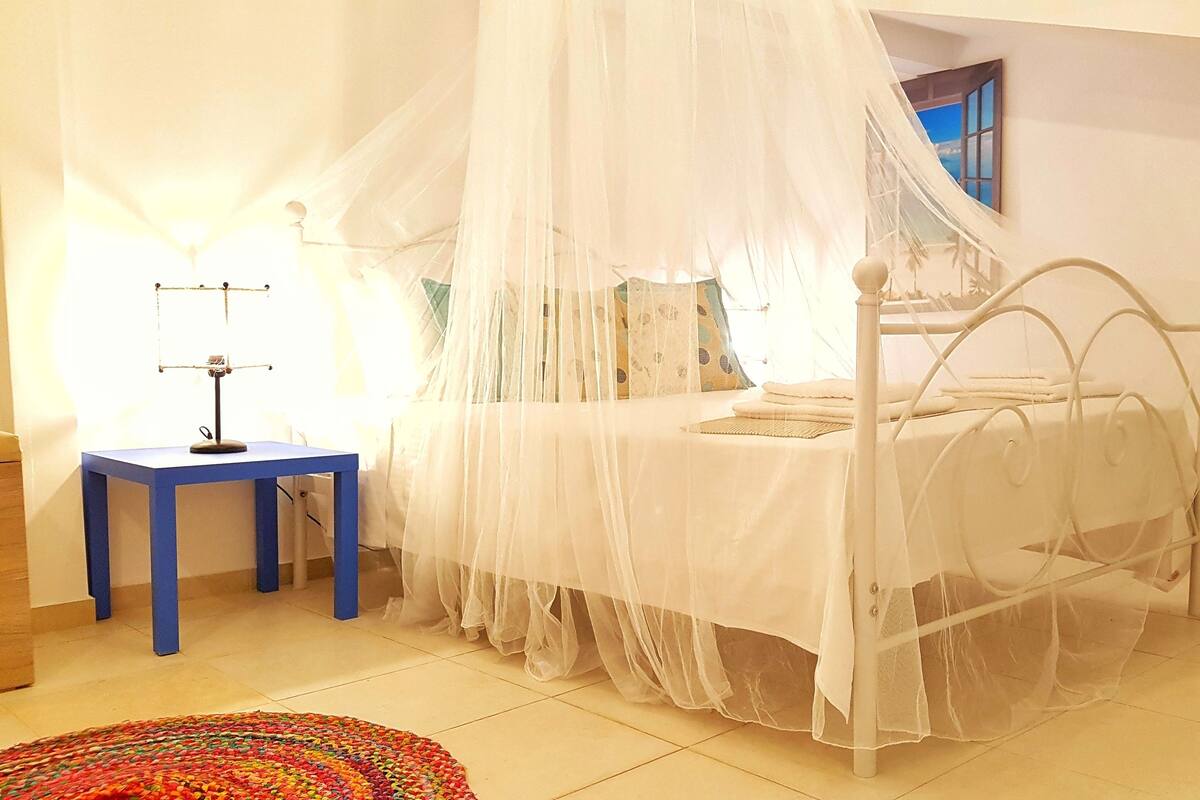 Une chambre à coucher avec un lit placé sous un baldaquin blanc, une petite table avec une lampe, une chaise et un tapis coloré sur le sol.