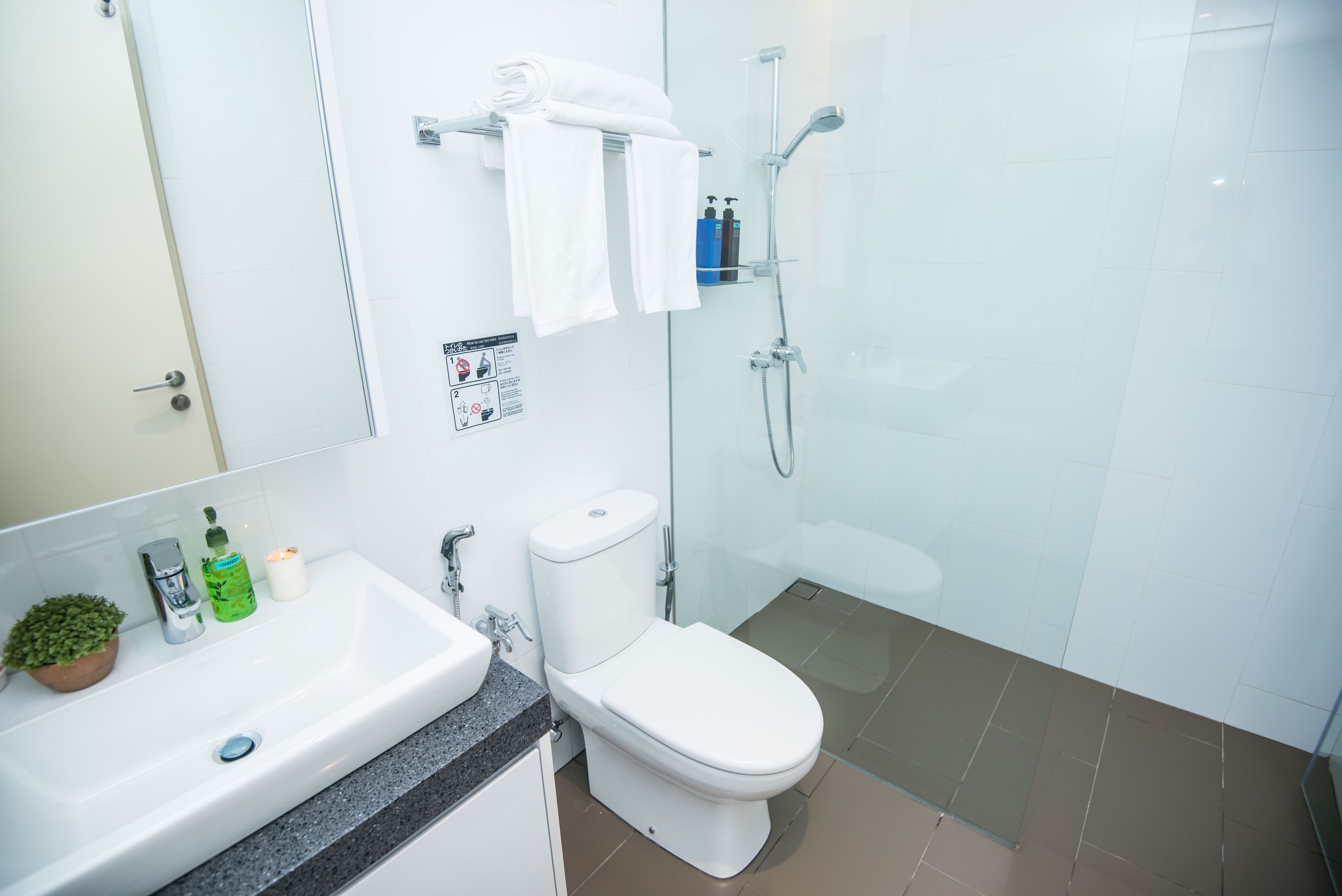 A modern bathroom with a white sink, toilet, and shower. There is a towel rack with folded towels, a small potted plant, and toiletries on the sink.