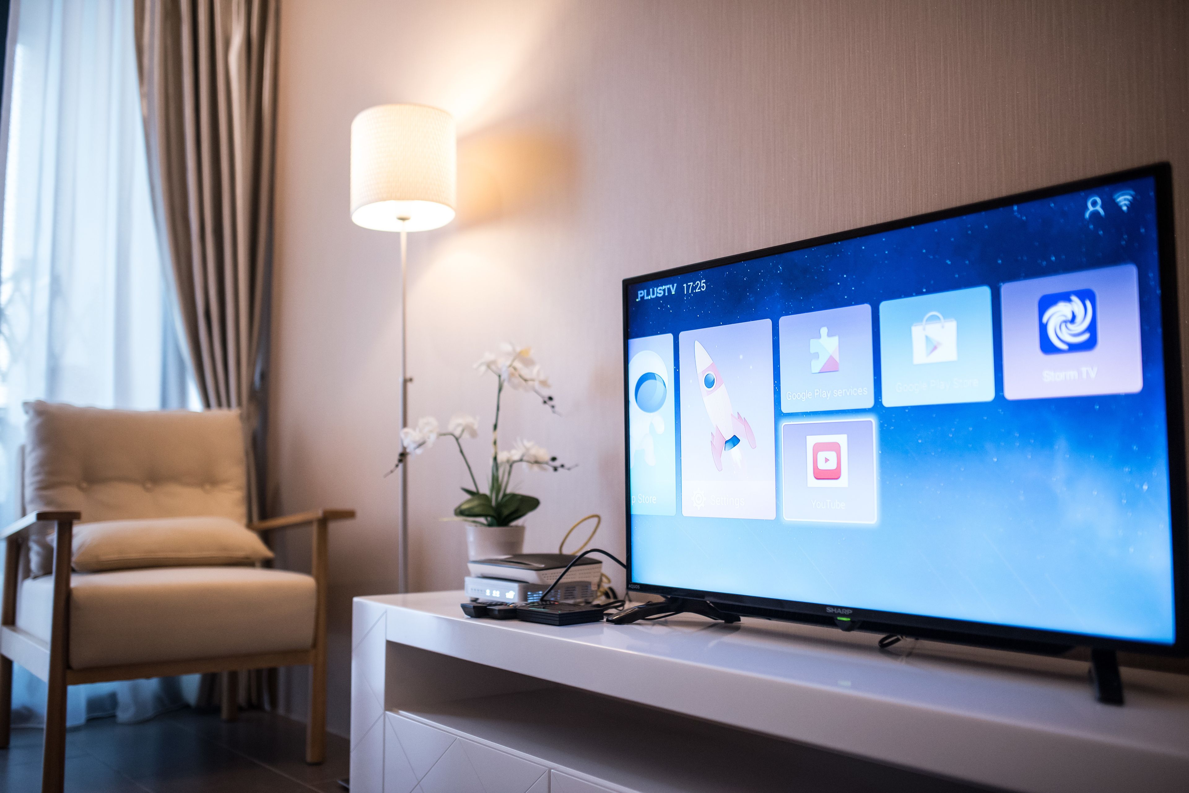 Un salón moderno con un televisor de pantalla plana sobre un soporte blanco, un sillón beige, una lámpara de pie y una pequeña planta en maceta.