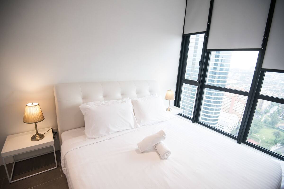 A hotel room with a large bed, two bedside lamps, and a view of the city through floor-to-ceiling windows.