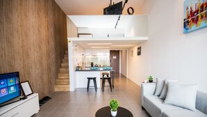 Uma sala de estar moderna com televisão de tela plana, uma mesa de centro com uma planta, um sofá cinza e uma área de cozinha ao fundo.
