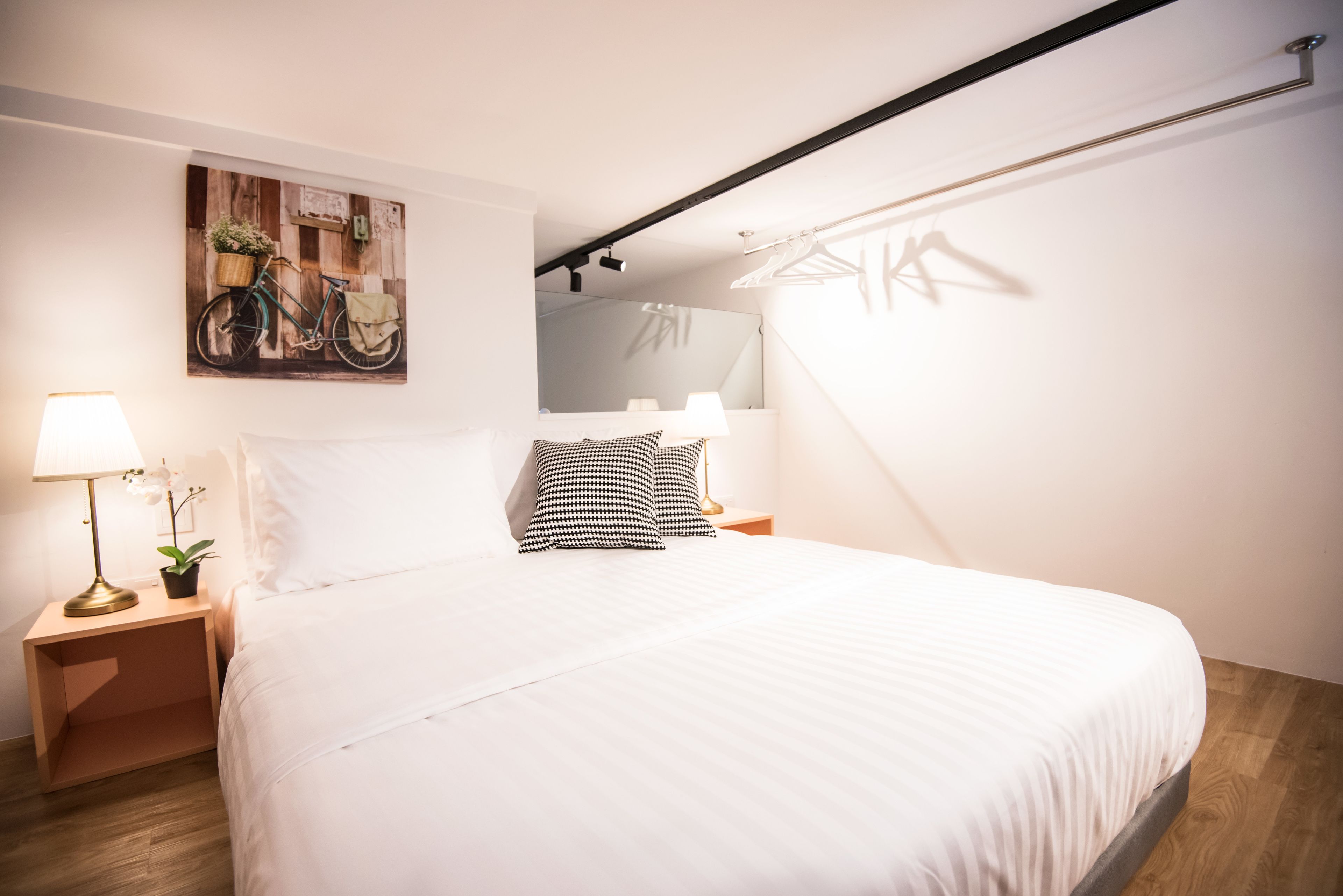 A neatly made bed with white linens, a bedside lamp, a small plant, and a wall-mounted painting of a bicycle.