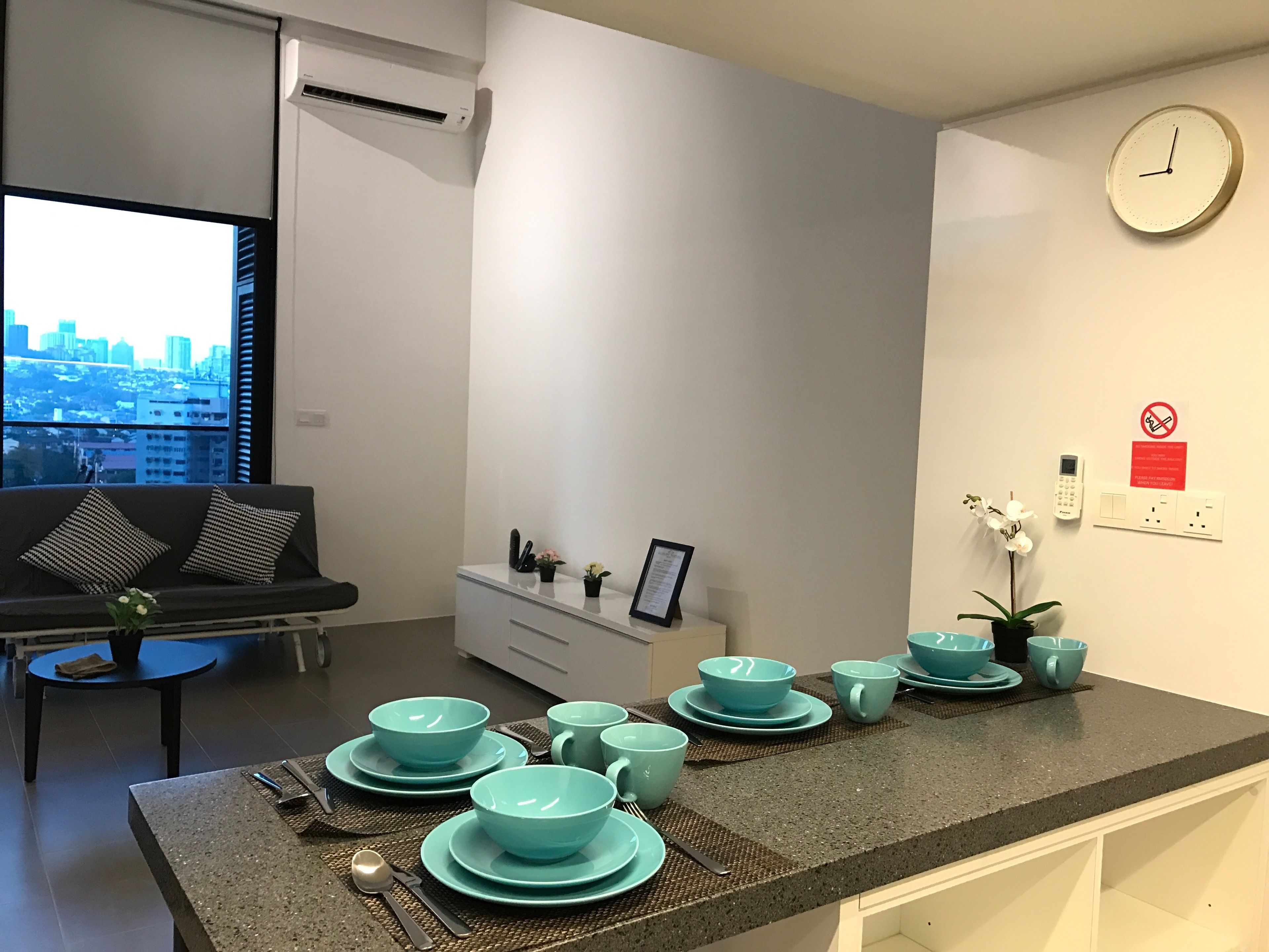 A modern kitchen with teal-colored bowls and plates on the counter, a sofa with patterned cushions, a city view through the window, and a wall-mounted clock.