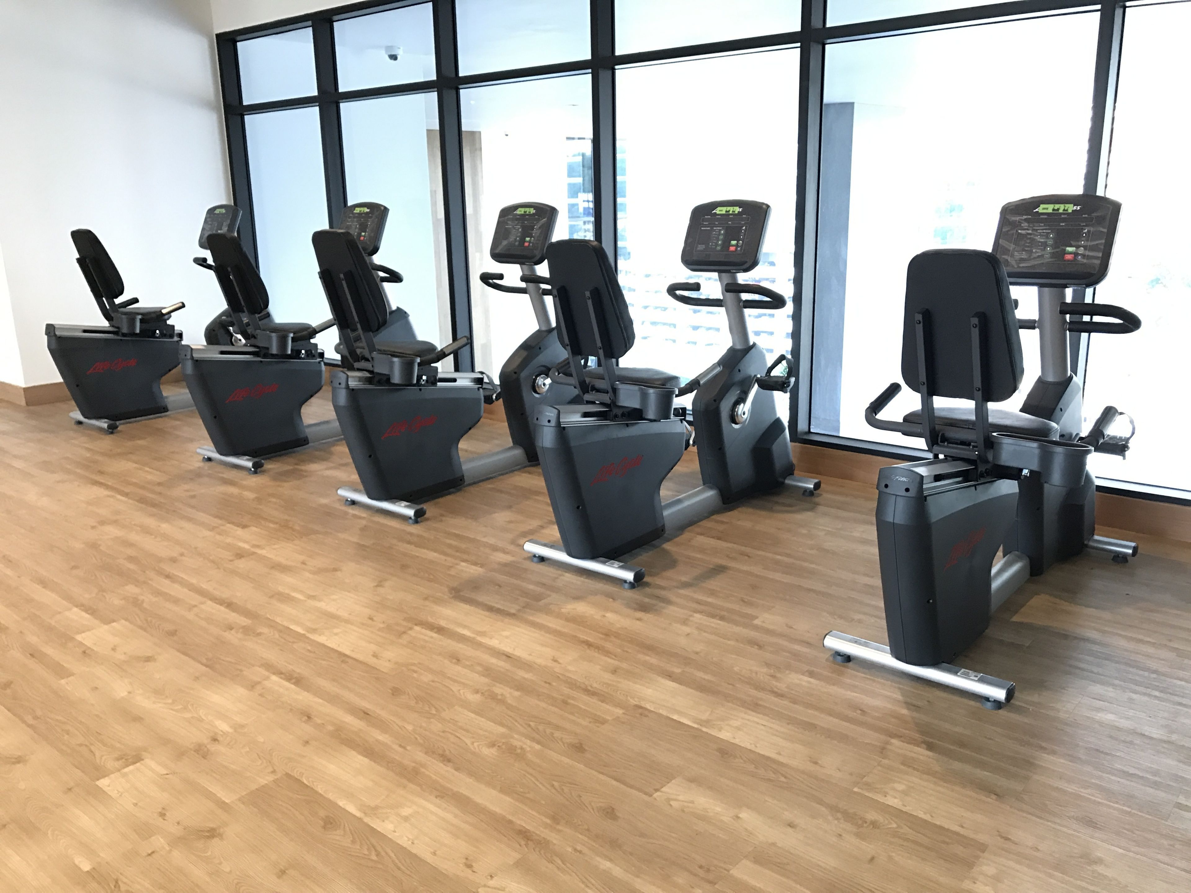 A row of stationary bikes in a gym with large windows and wooden flooring.