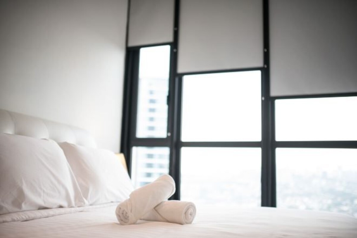 Ein ordentlich bezogenes Bett mit weißen Leinen und einem zusammengerollten Handtuch, große Fenster mit Jalousien und Blick auf die Stadt.