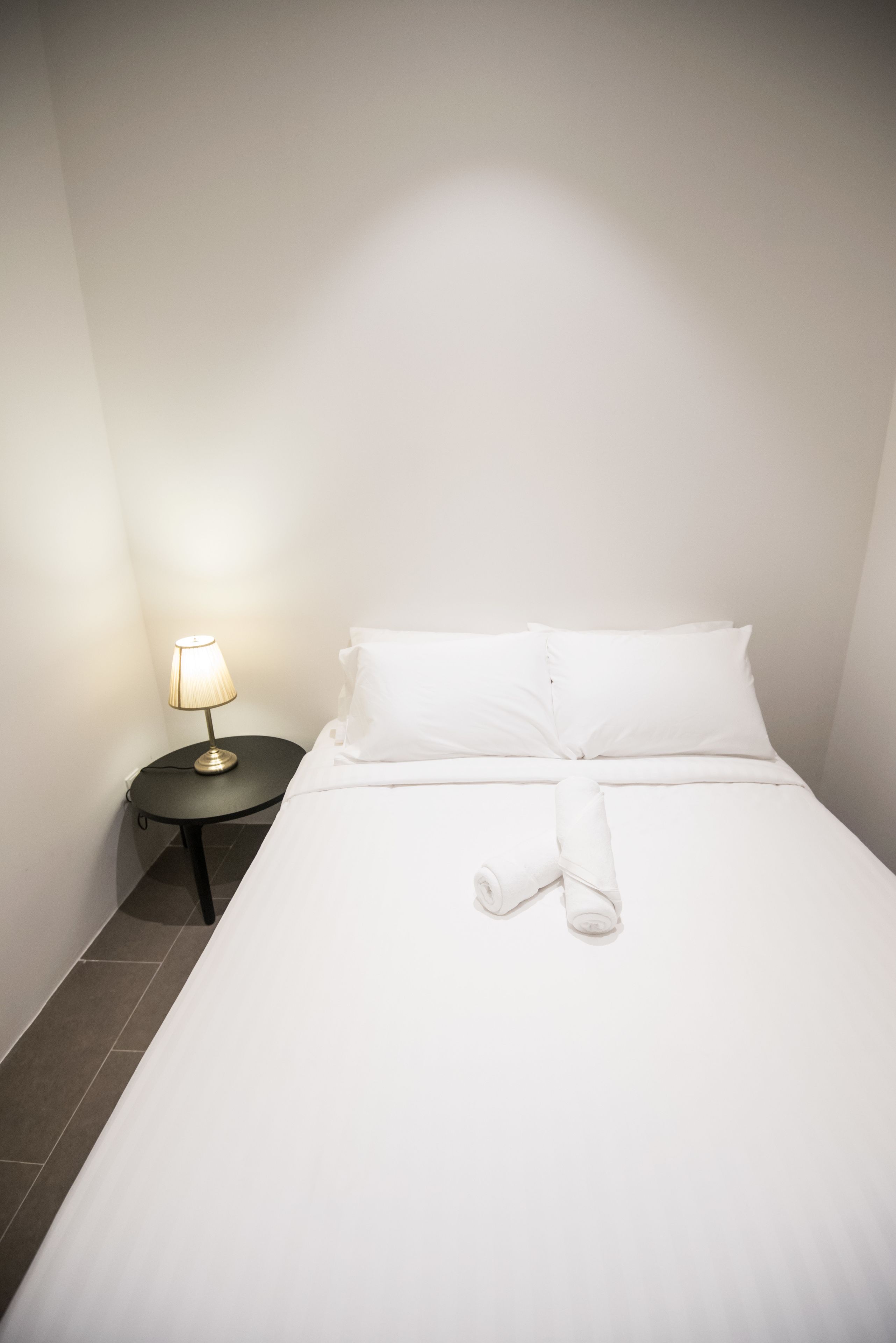 A neatly made bed with white linens and a rolled towel, a small round table with a lamp, and a plain white wall.